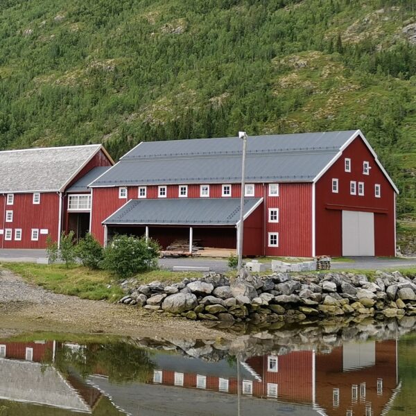 Stor rød trebygning med hvite vinduer. Hav i forgrunn. Grønn fjellside bak