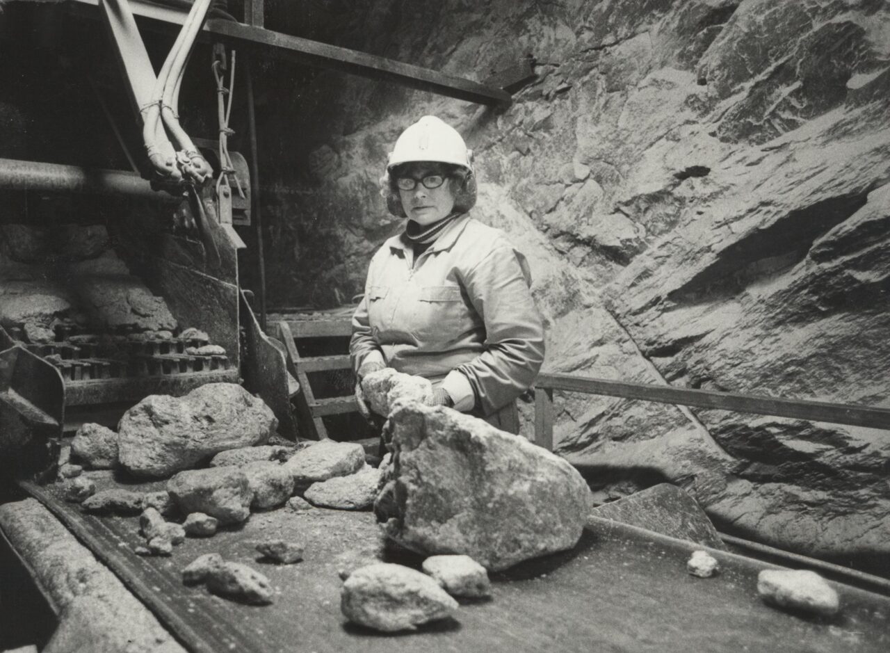 Dame med briller og hjelm står bak et bord med steiner. Sorthvitt foto