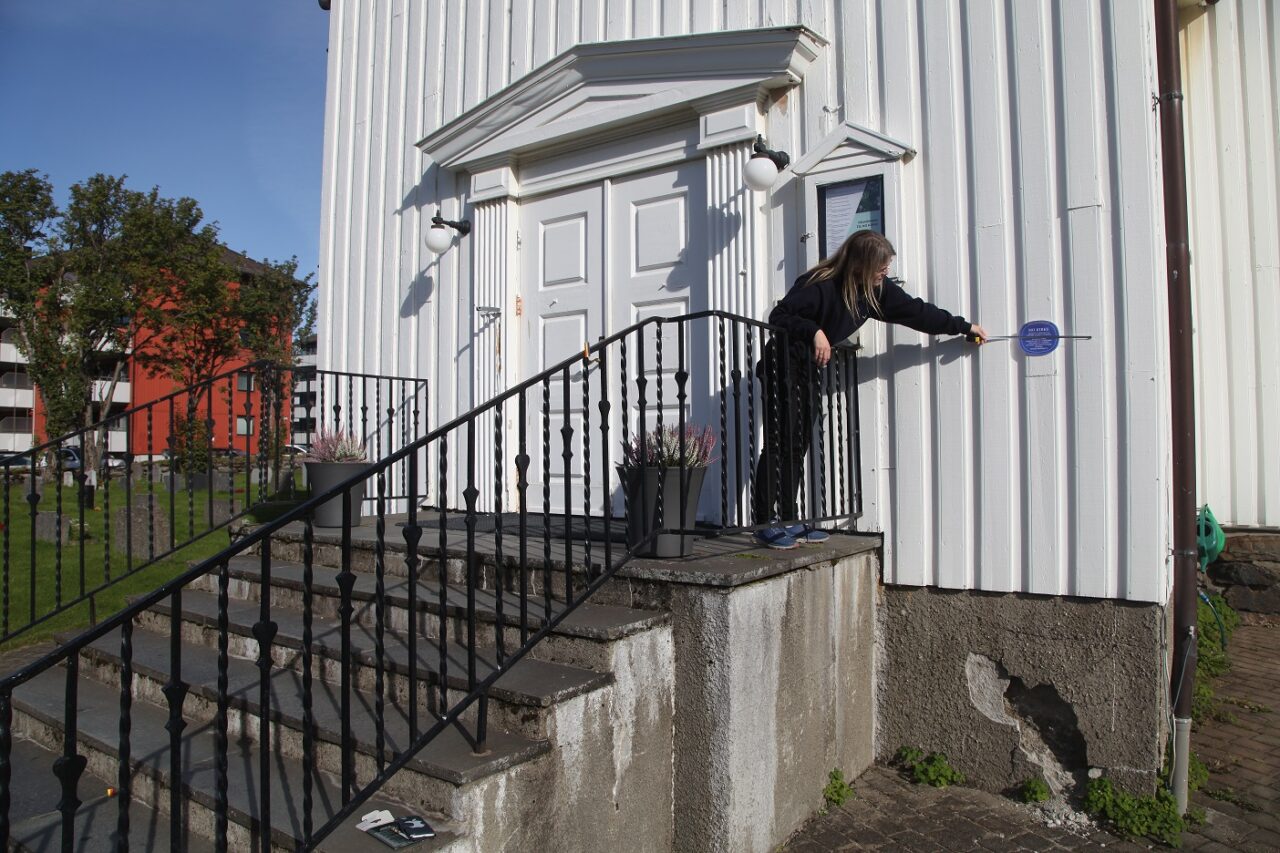 Dame i sort står på ei trapp og måler et blått skilt på hvit husvegg. Foto