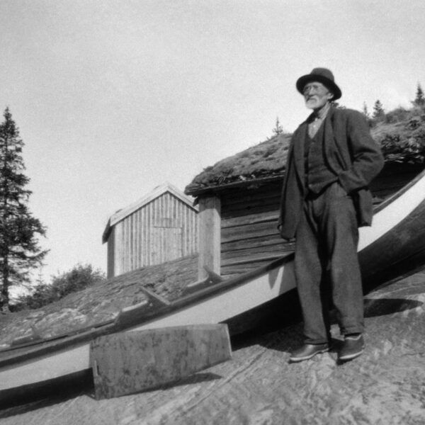 En mann med hatt og briller står ved siden av en trebåt på et berg. To trehus i bakgrunn