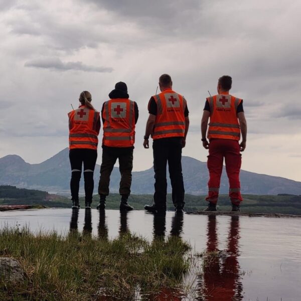 Fire personer med oransje refleksvester står med ryggen til fotografen. Vann i forgunn, fjell i bakgrunn