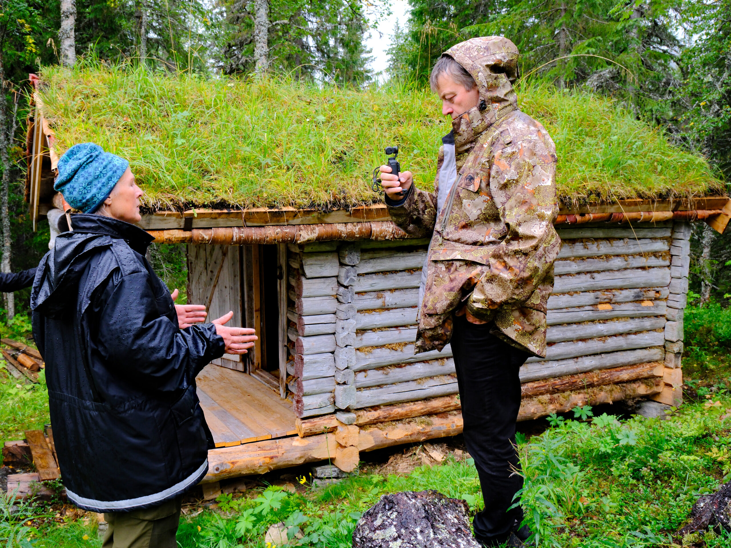 Et laftet bur med to mennesker fremfor. Skog omkring