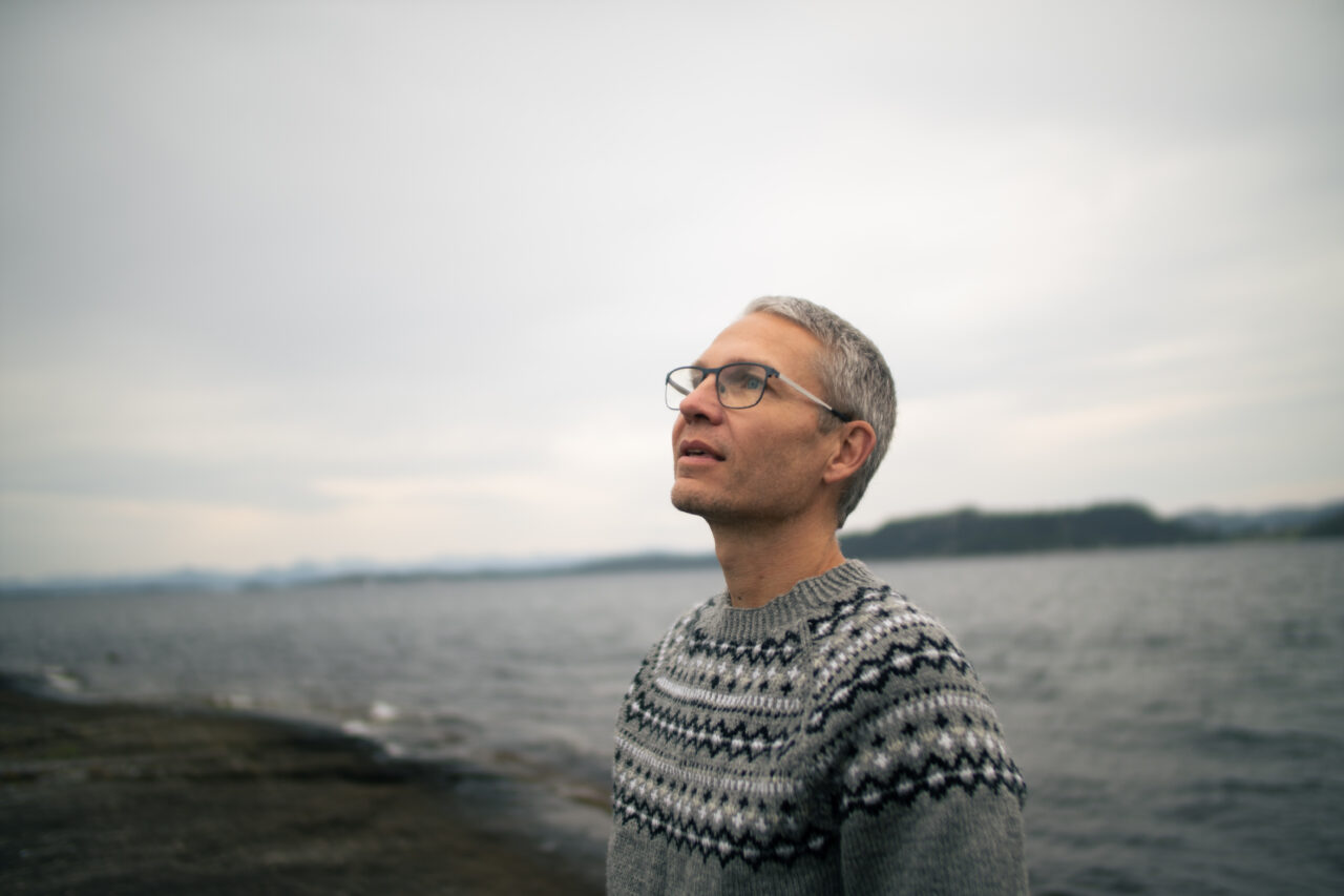 Person med briller og mønstret strikkegenser står ved vannkanten, med landformer i bakgrunnen under overskyet himmel. Foto.
