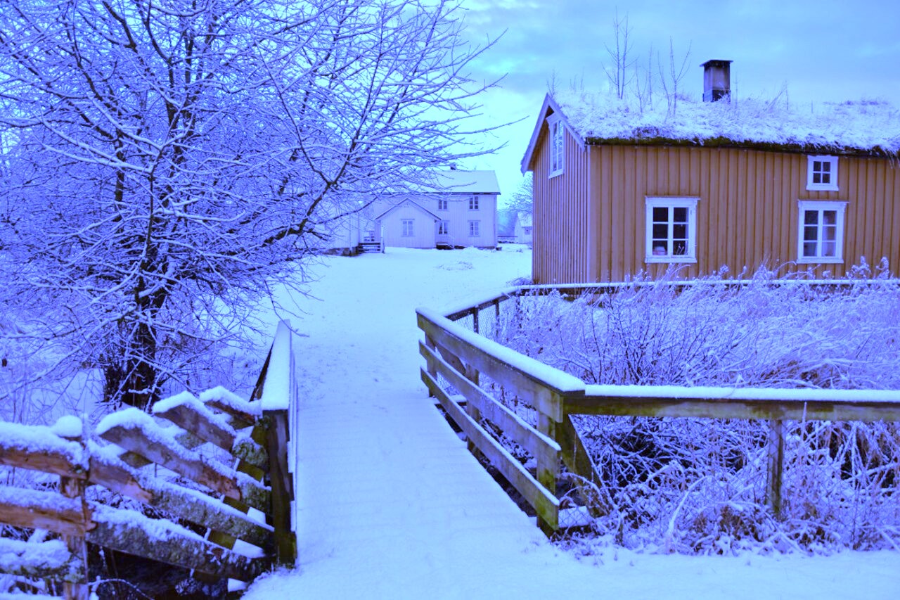 Snødekt vinterlandskap med trebro som leder til et hus med hvite vinduskarmer og snødekt tak.