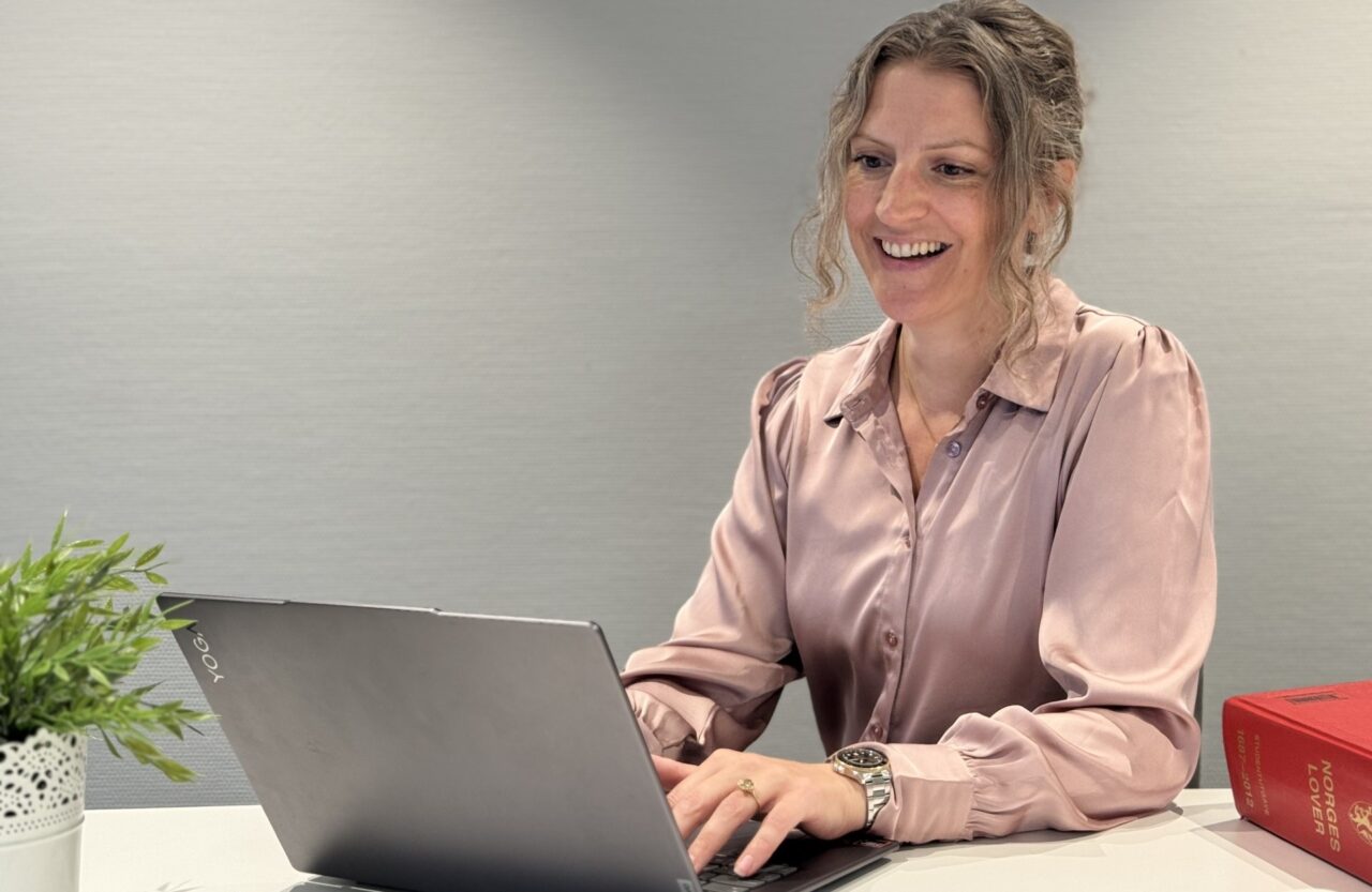 Dame i med støvrosa silkebluse sitter ved en pult med laptop foran seg. Foto