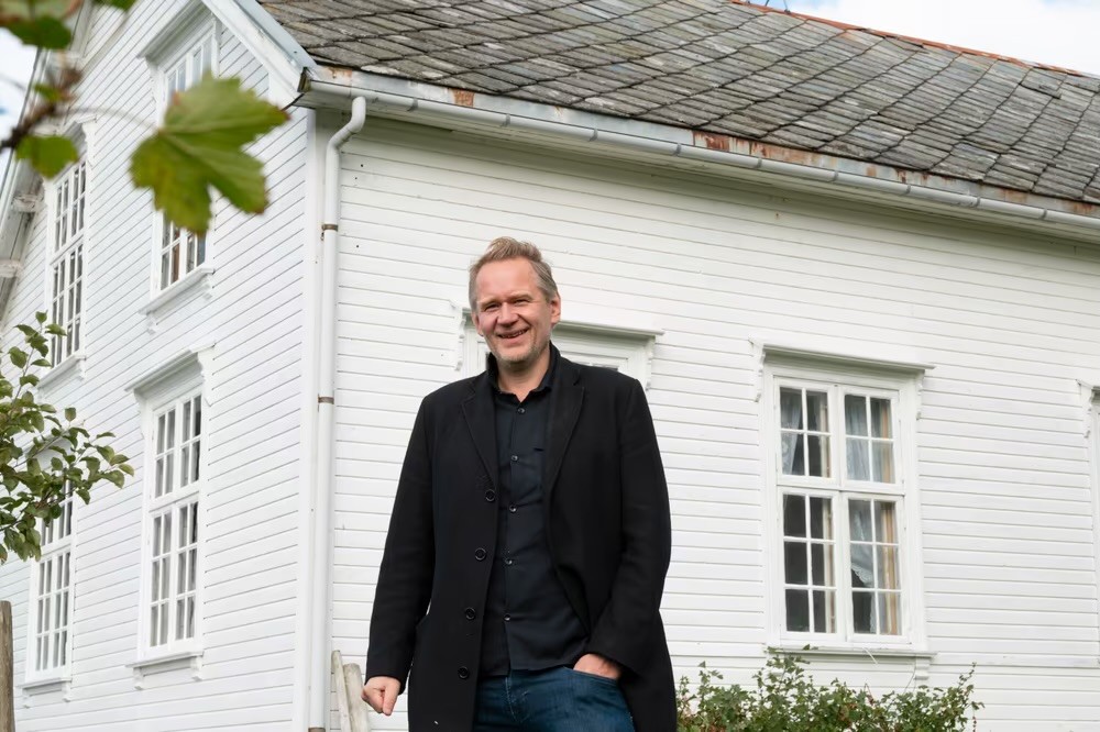 Smilende person i mørk jakke og jeans foran hvitt trehus med takstein og sprossevinduer, omgitt av grønne blader