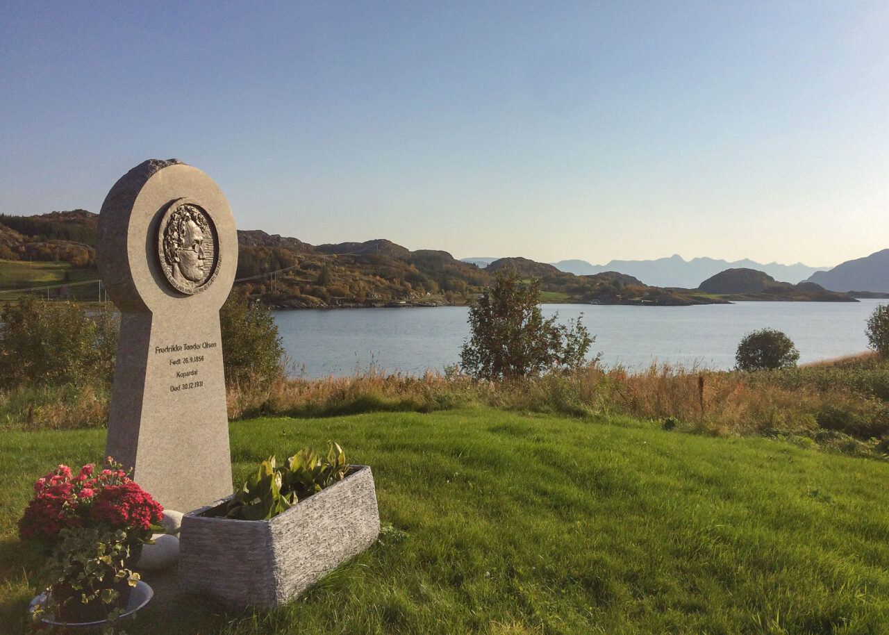"Minnesmerke for Fredrikke Olsen Tønder. Plassert ved sjøen med fjell i bakgrunnen. Blomster ved foten av steinen. Foto