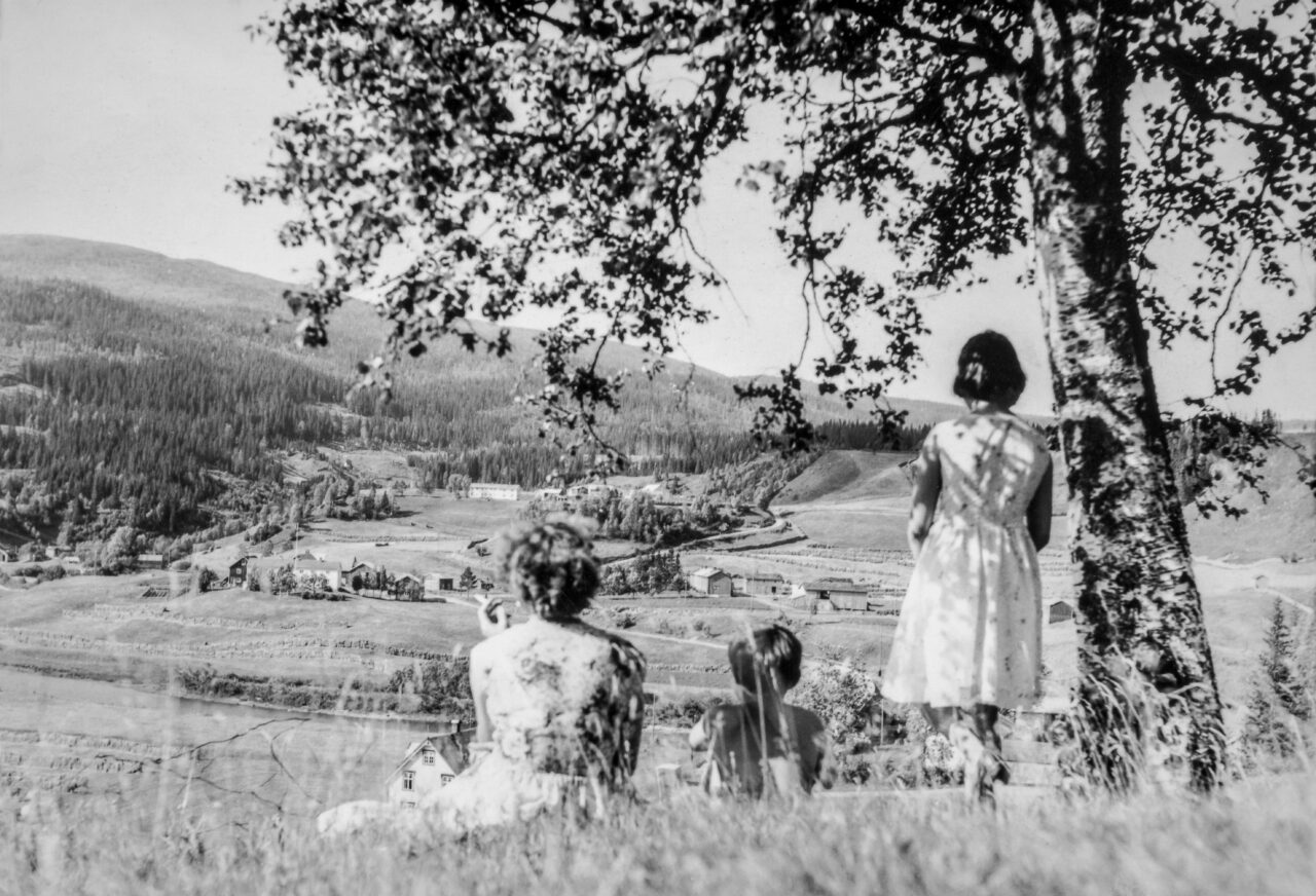 Tre personer sitter i gresset under et tre og ser utover en dal med gårder og skogkledde åser. Bildet er i svart-hvitt og gir et rolig landskapsperspektiv.