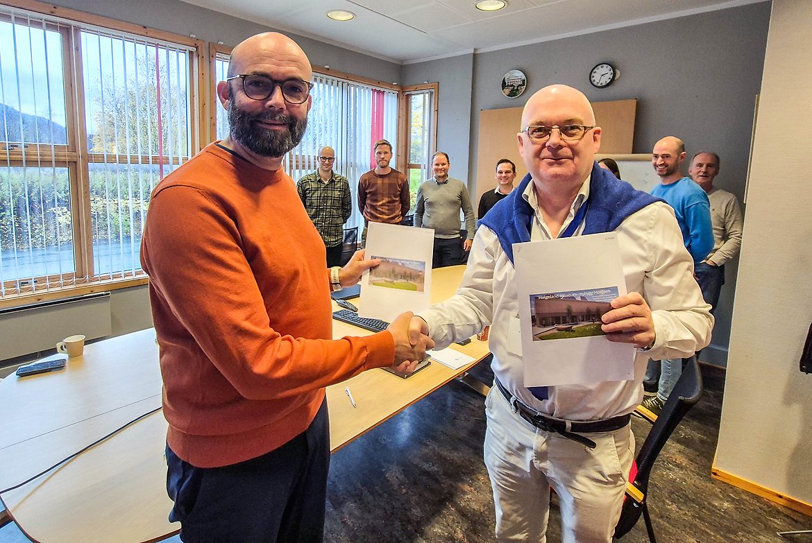To personer håndhilser i et møterom mens de holder bilder av en bygning. Flere personer står i bakgrunnen langs veggen. På bordet ligger dokumenter og en kaffekopp.