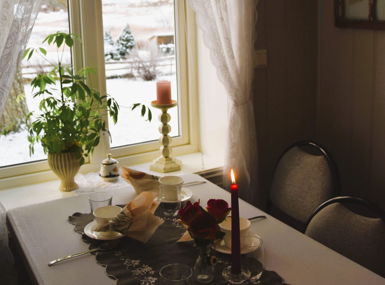 Dekket bord med kaffekopper, levende lys og blomster ved et vindu med utsikt til snødekt landskap.