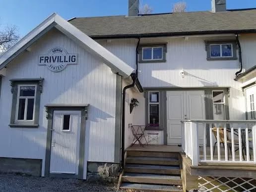 Hvit trebygning med skiltet ‘Sømna Frivilligsentral’ på veggen, inngangsdør med trapp og liten terrasse med bord og stoler