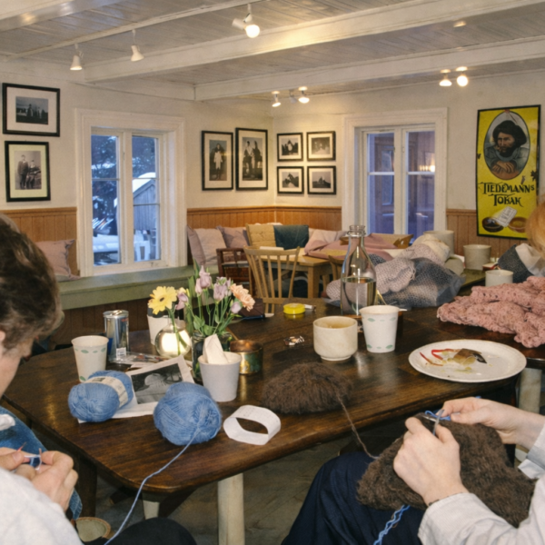 Personer sitter rundt et bord og arbeider med garn og strikketøy. På bordet ligger nøster, kopper, en vase med blomster og en tallerken med matrester. Rommet har flere vinduer, veggdekor og innrammede bilder.
