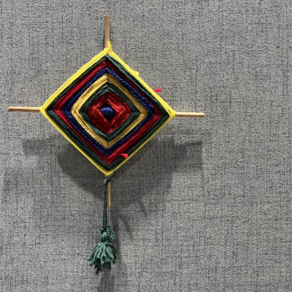 Fargerikt vevd ornament laget av garn i flere lag festet på to kryssede trepinner. Formen er diamantformet med rødt, blått, gult og grønt garn. En grønn dusk henger fra nederste pinne mot en grå bakgrunn.