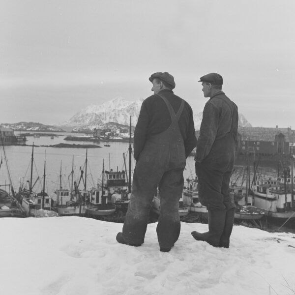 To fiskere i arbeidsklær står på en snødekt høyde og ser utover en havn fylt med fiskebåter. I bakgrunnen reiser et spist, snødekt fjell seg over kystlandskapet i dette historiske sort-hvitt-motivet, foto.