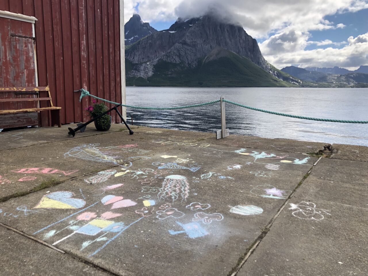 Fargerike kritttegninger av sjødyr og figurer på en betongkai ved siden av en rød sjøbu. I bakgrunnen ser man en rolig fjord og et høyt, spisst fjell med skyer rundt toppen, foto.