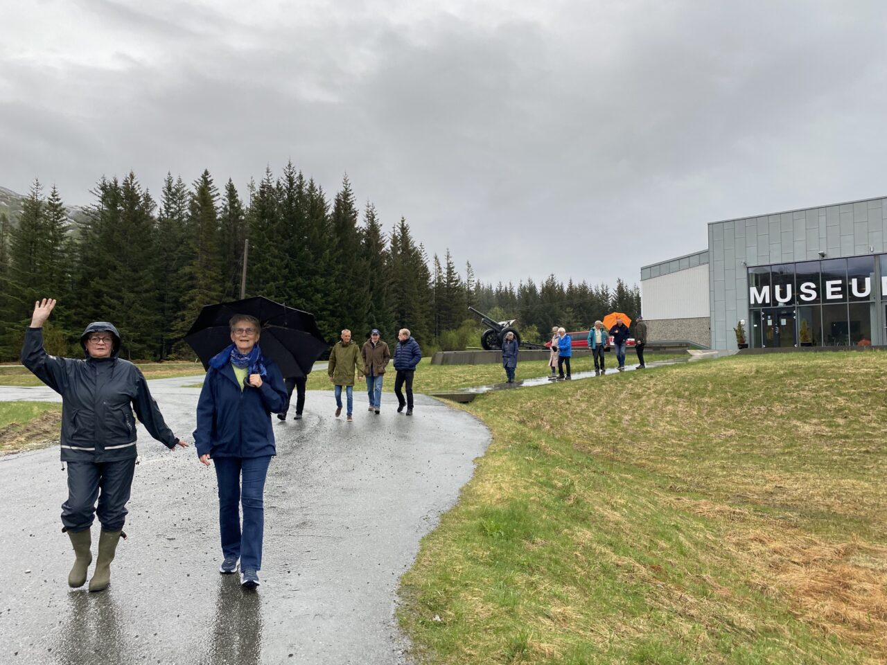 To kvinner i turklær går fremst, mens resten av følget kommer etter fra skogkanten og forbi en utstilt kanon, foto.