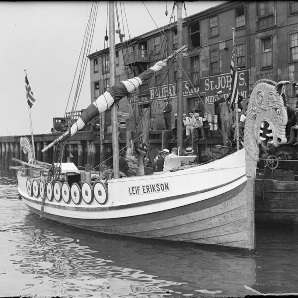 Vikingskipet «Leif Erikson» ligger til kai foran en eldre trebygning med reklameskilt. Skipet har et utskåret dragehode i baugen, skjold langs siden og både norsk og amerikansk flagg. Foto.