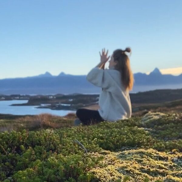 En person sitter i meditasjon på en høyde dekket av mose og lyng i forgrunnen. Personen har hendene samlet foran ansiktet og er vendt mot et åpent kystlandskap med en fjord og en markant, taggete fjellrekke i bakgrunnen under en klar, blå himmel.