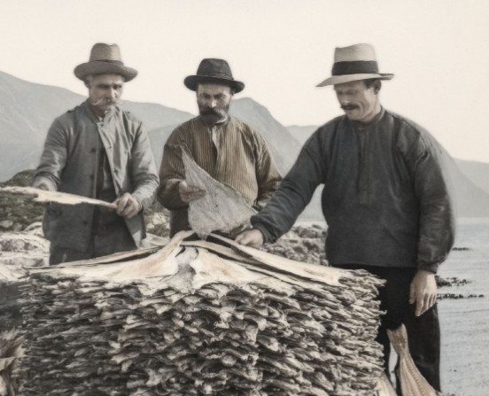 Tre menn i hatter og historiske klær står foran en stor stabel med tørket tørrfisk ved en fjord med fjell. Sorthvitt-foto.