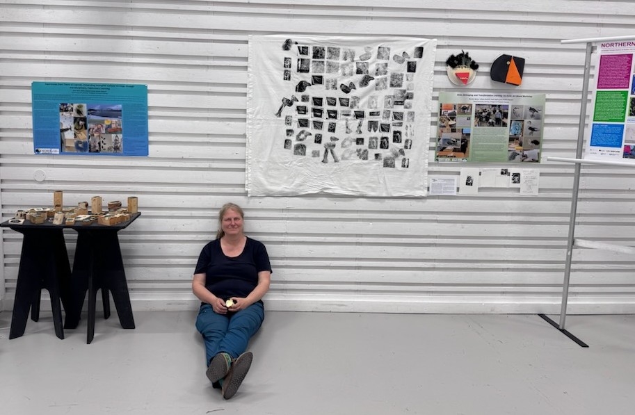 Person sitter på gulvet foran en vegg med en oppslagstavle med fotografier, bilder og kunstverk i et utstillings- eller gallelokale. Foto