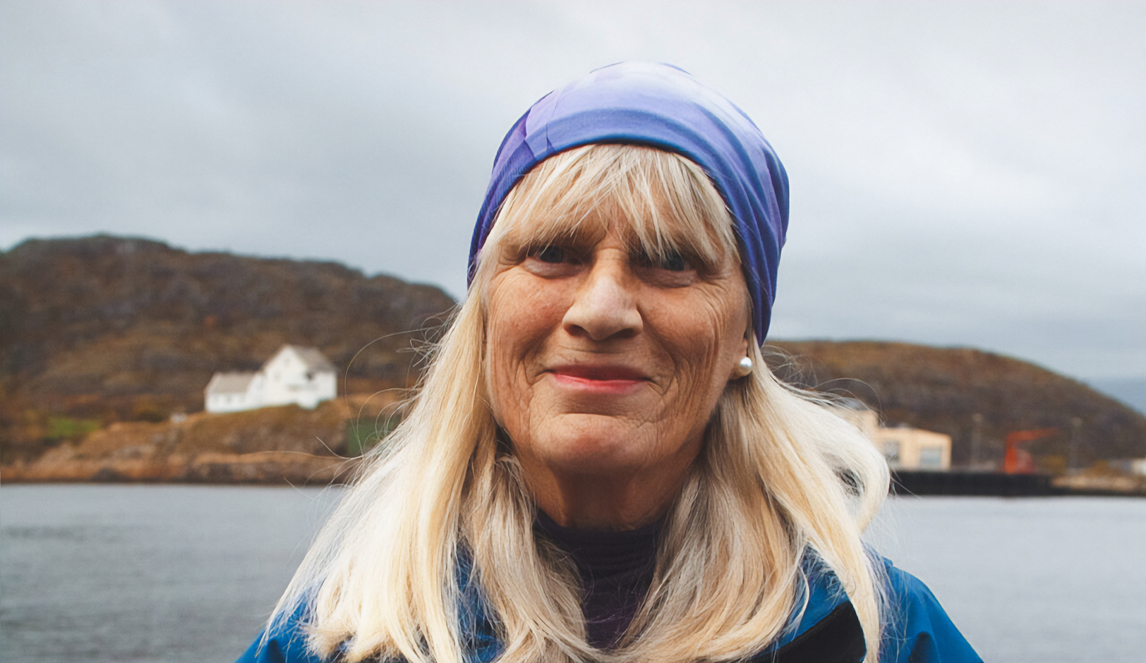Portrett av en person med lyst hår og blått hodeplagg ved vannkanten, med hus og lave åser i bakgrunnen.