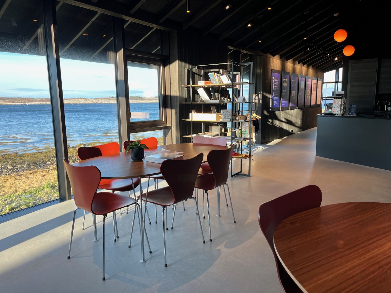 Foto. Interiør i Vevelstad besøkssenter med røde og oransje skallstoler rundt ovale trebord, store vinduer med utsikt mot sjø og holmer, og lilla utstillingsplakater langs mørk trevegg.