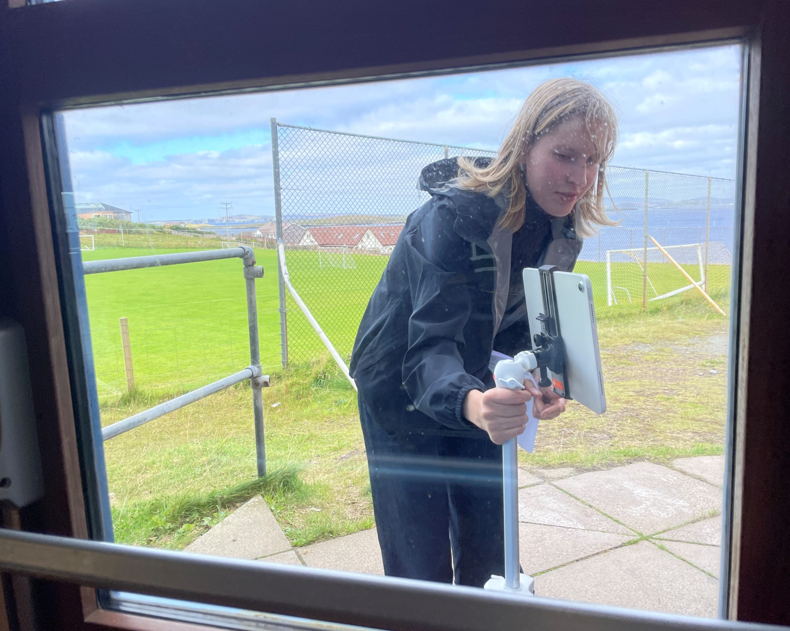 Kvinne vasker et vindu utenfra med nal og skrape, sett fra innsiden gjennom glasset. Foto
